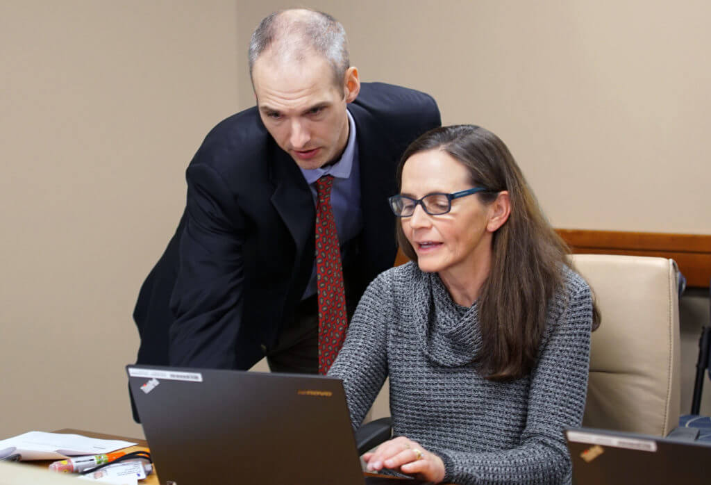 Schuyler Jones, left, stands behind Patty McAdams, who is seated at a desk. They both look at a computer together. 