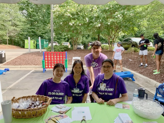 Three people in purple shirts sit at a table in a parking lot at an event.