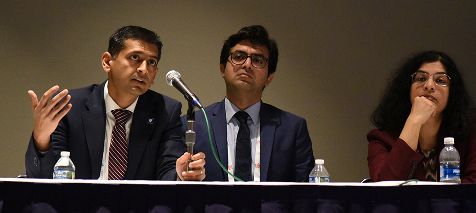 Nishant Shah speaks into a microphone as two panel participants look on thoughtfully.