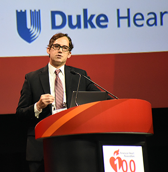 DCRI's Jonathan Piccini presents his thoughts on the PROMPT-AF trial while standing at a podium on the main stage of the AHA Scientific Sessions. The Duke Heart Center logo appears above his head in the background on the screen behind him.