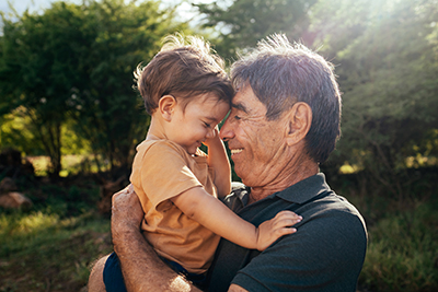 A grandparent smiles as he embraces his young grandchild while standing outside on a sunny day.