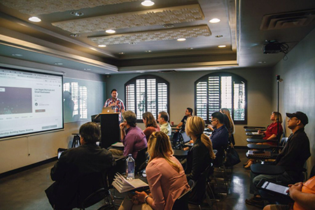 Person presenting at the front of a conference room while attendees sit and take notes, with a large screen displaying slides on the wall.