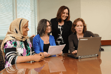 Four women gathered around a laptop in a conference room, reviewing content on the screen. One woman holds a printed document while another points at the laptop.