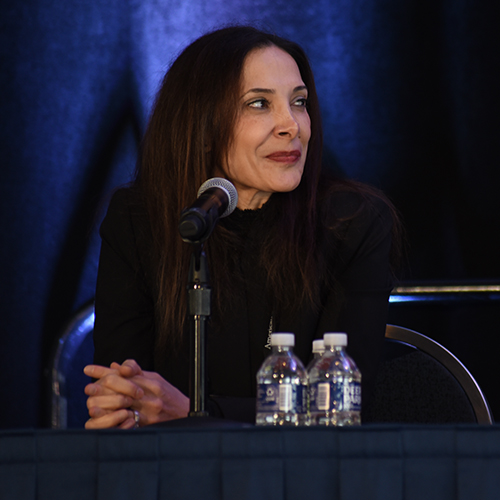 Sana Al-Khatib sits at a panelists table during a recent conference, glancing to her right as she smiles at the presenter. The background is purple, and her hands are folded on the table in front of the microphone stand resting in front of her.