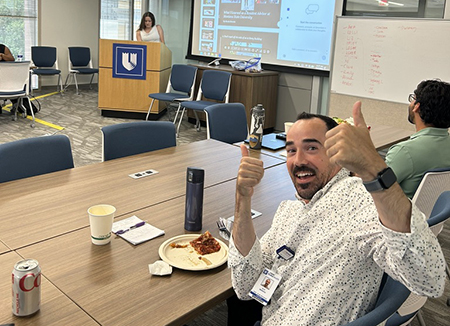 Conference room scene showing a participant seated at a large meeting table giving two thumbs up toward the camera. A plate with a slice of pizza, a beverage can, a coffee cup, and a water bottle sit on the table nearby. In the background, a presenter stands at a podium bearing the Duke shield logo while presentation slides are displayed on a screen. Additional attendees are seated around the room, and a whiteboard with handwritten notes is visible near the front.