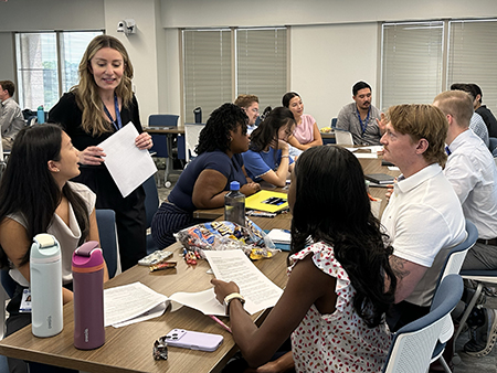 A facilitator stands beside seated participants in a classroom-style conference room, holding a document while speaking with attendees. Several small groups are gathered around tables reviewing printed materials and taking part in discussion. 