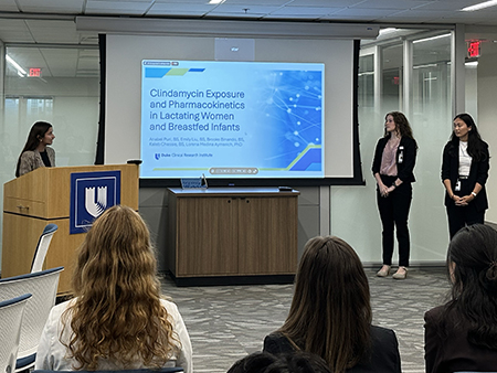 Three presenters stand at the front of a conference room beside a projected slide titled “Clindamycin Exposure and Pharmacokinetics in Lactating Women and Breastfed Infants.” Audience members are seated and facing the presentation.
