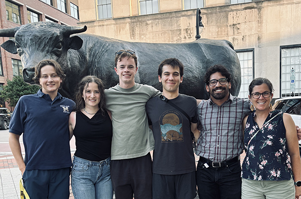 Group photo of six people standing outdoors in front of a large bronze bull statue in an urban plaza.