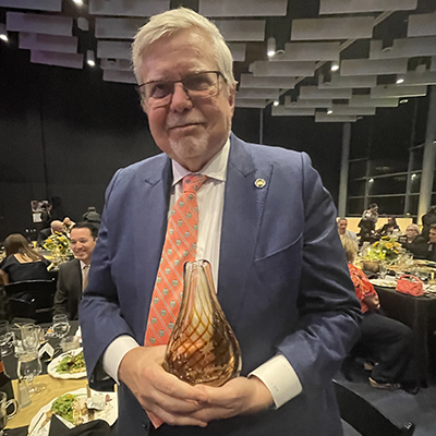 Frank Rockhold poses in a blue suit and patterned orange tie holds a glass award at a banquet event with tables and attendees in the background.