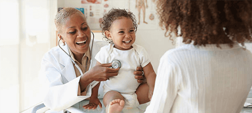 A smiling doctor uses her stethoscope to listen to the heart of a happy baby who is smiling at their caregiver. The caregiver has their back turned to the camera.