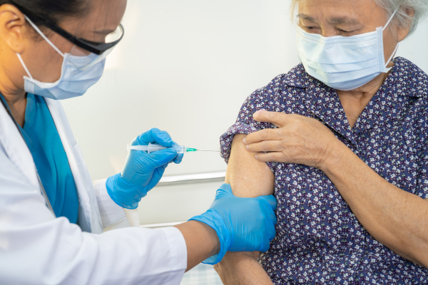 A clinician injects a vaccine into the upper arm of an elderly woman. Both individuals are wearing face masks.