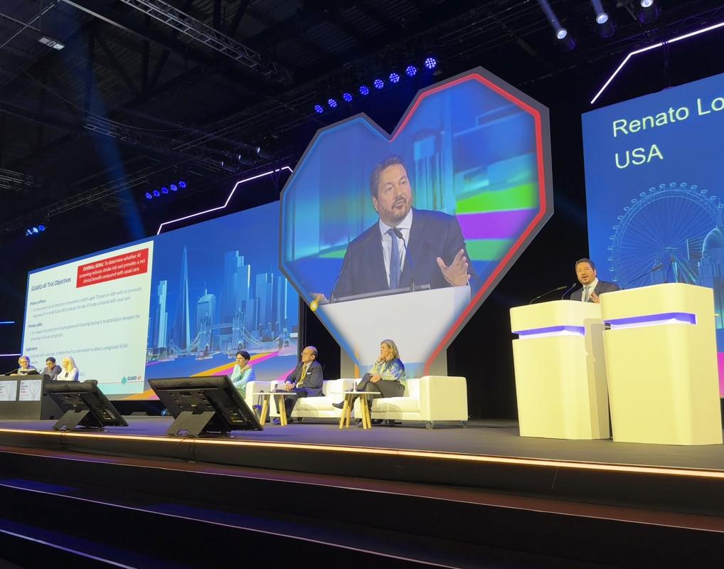 Renato Lopes, MD, PhD, speaks at a podium. Behind him, his image is projected on a heart-shaped screen, with panelists sitting nearby. 