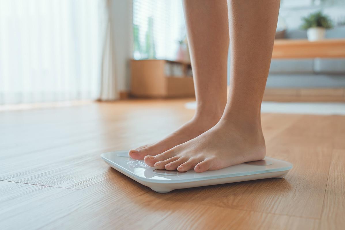 A woman's feet are seen standing on a white scale sat on a hardwood floor as she measures her weight.