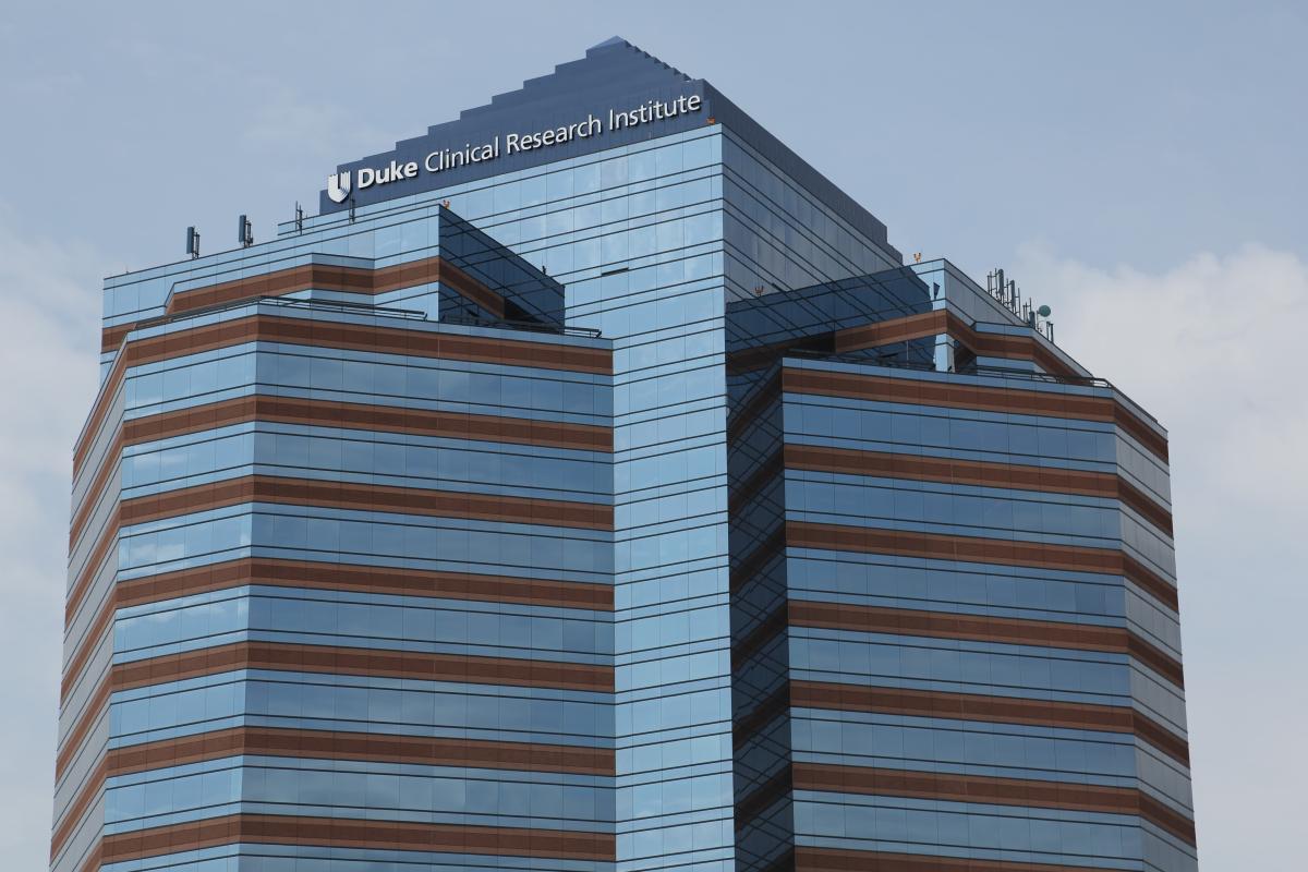 A photo of The Grove Durham building, in downtown Durham. The building is tall and blue, with lots of windows and a pyramidal top. It says Duke Clinical Research Institute at the top next to the Duke shield. 