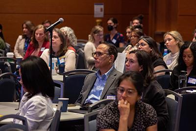 Audience seated at round tables in a conference room, listening to a speaker; a standing microphone is positioned among attendees, and a presentation screen is visible at the front.