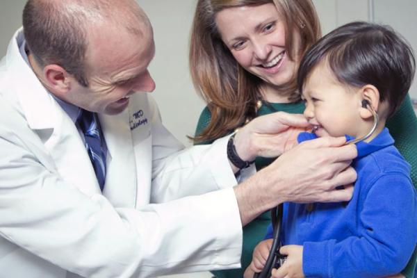 A doctor interacts with a young patient sitting on their mother's lap.