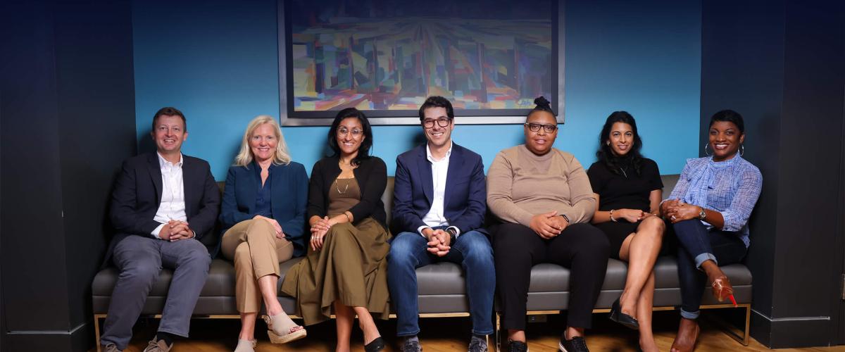 The team of seven i-Cubed leaders sit on a couch, smiling against a deep blue background.