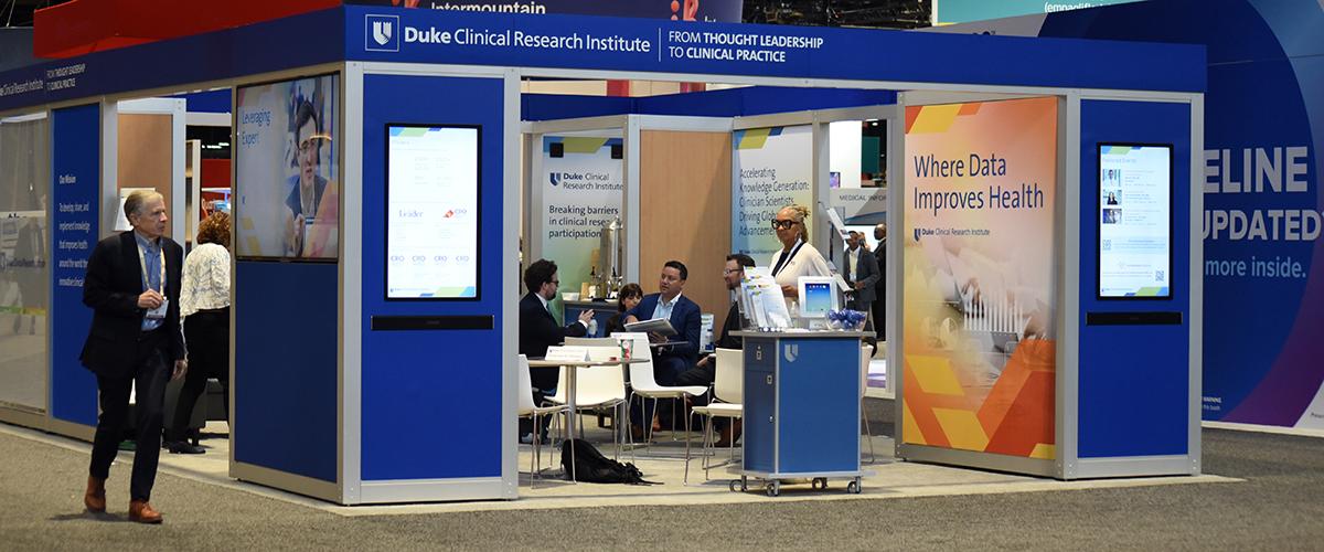 The DCRI Booth is shown with Chris Granger walking past on the left hand side, and multiple meetings taking place inside the Duke Blue structure in the McCormick Place conference exhibits hall in Chicago.