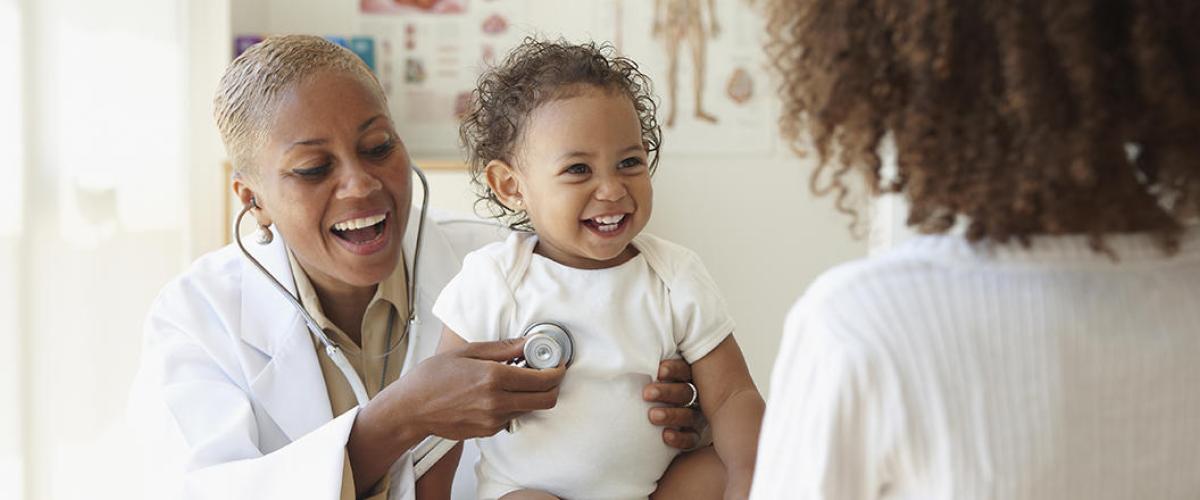 A doctor laughs as they hold a stethoscope to a laughing baby's chest as the mother looks on with her back to the camera.