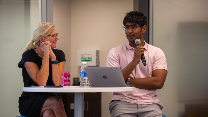 A woman sits at a small table speaking with a man holding a microphone. 