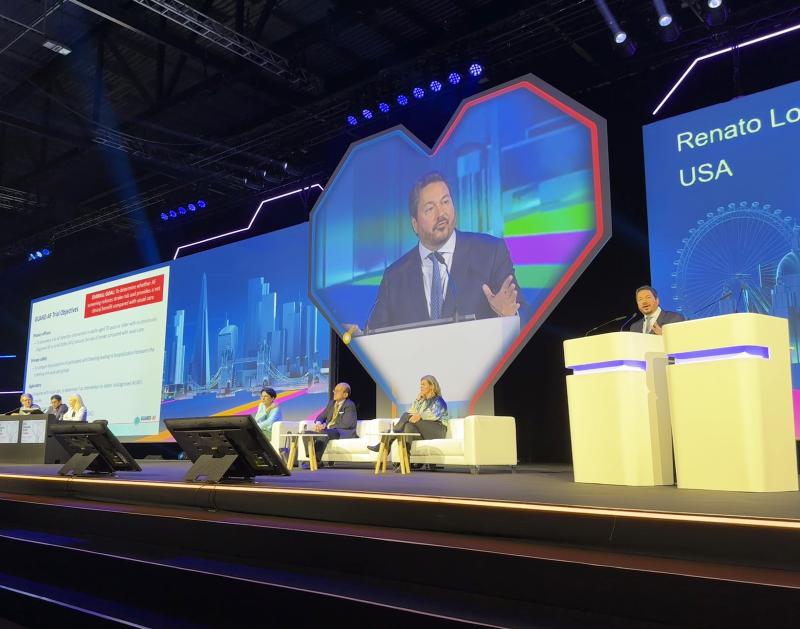 Renato Lopes, MD, PhD, speaks at a podium. Behind him, his image is projected on a heart-shaped screen, with panelists sitting nearby.