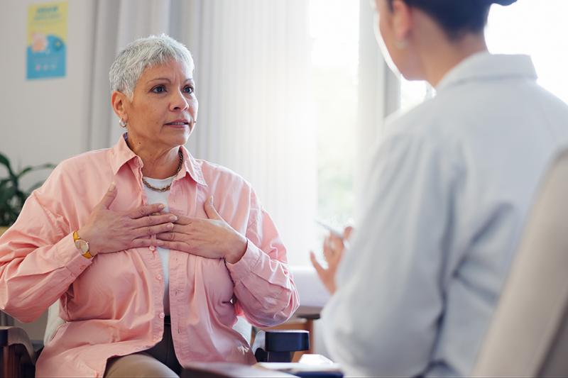 A woman speaks to a healthcare provider in a white coat while gesturing to her heart.