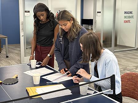 Three people stand around a table in a modern office, reviewing printed documents together. A table tennis net stretches across the foreground, and glass-walled meeting rooms are visible in the background. Text on a wall reads, “Imagine. Incubate. Ignite.”