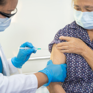 A clinician injects a vaccine into the upper arm of an elderly woman. Both individuals are wearing face masks.