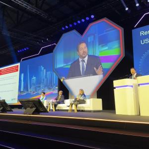 Renato Lopes, MD, PhD, speaks at a podium. Behind him, his image is projected on a heart-shaped screen, with panelists sitting nearby. 