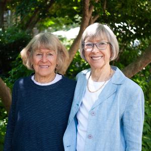 Two women stand together in front of a tree. 