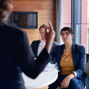 Two people sit in a meeting listening attentively to a third person whose back is turned to the viewer.