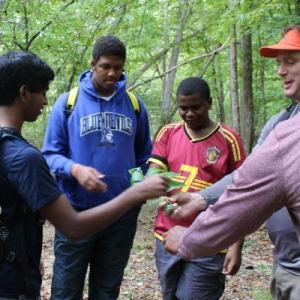 Three teens are seen smiling and interacting with two adult guides in a forest.